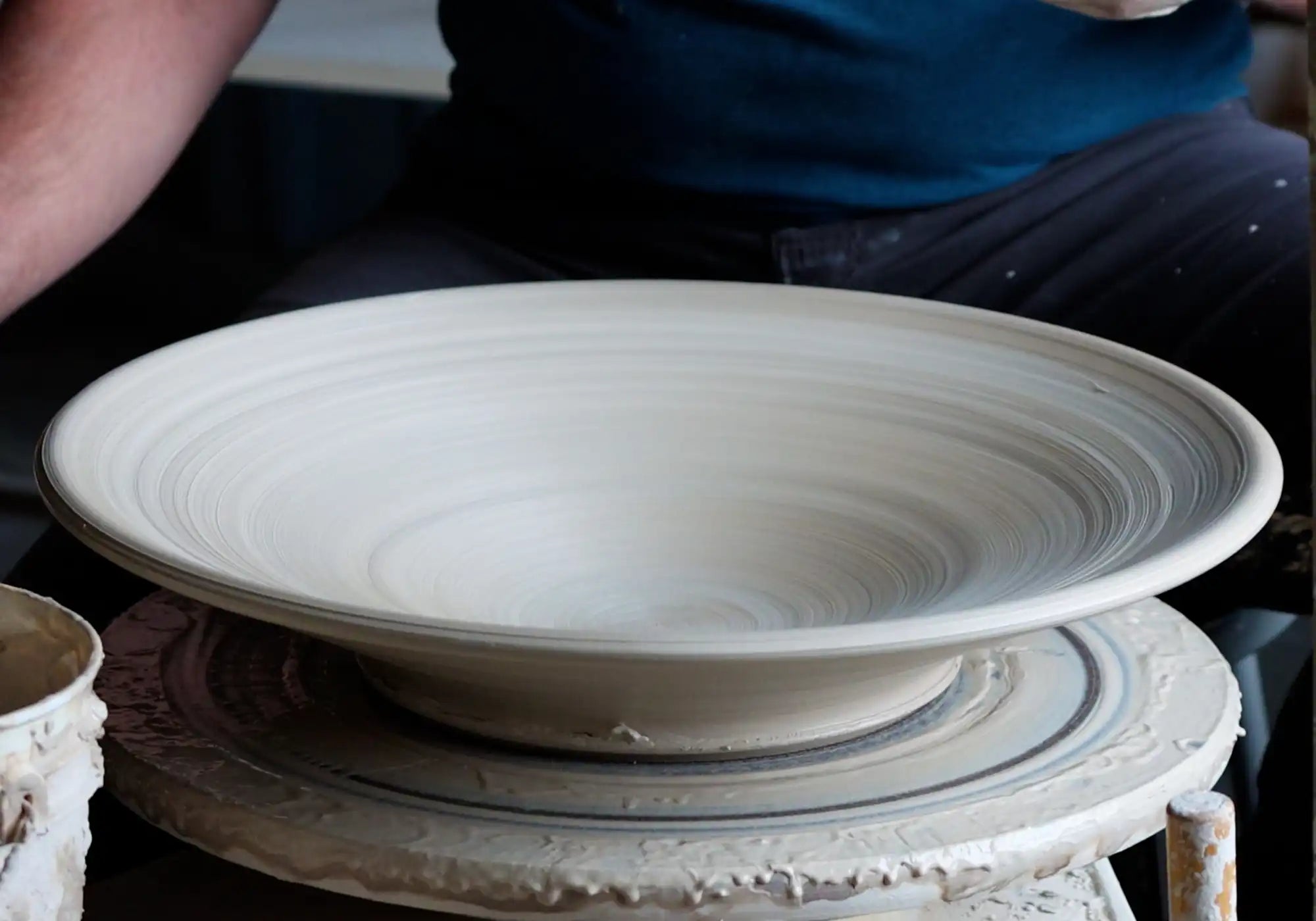 A clay bowl setting on a potter's wheel, freshly thrown by the pottery whose legs can be seen to either side of the potter's wheel.
