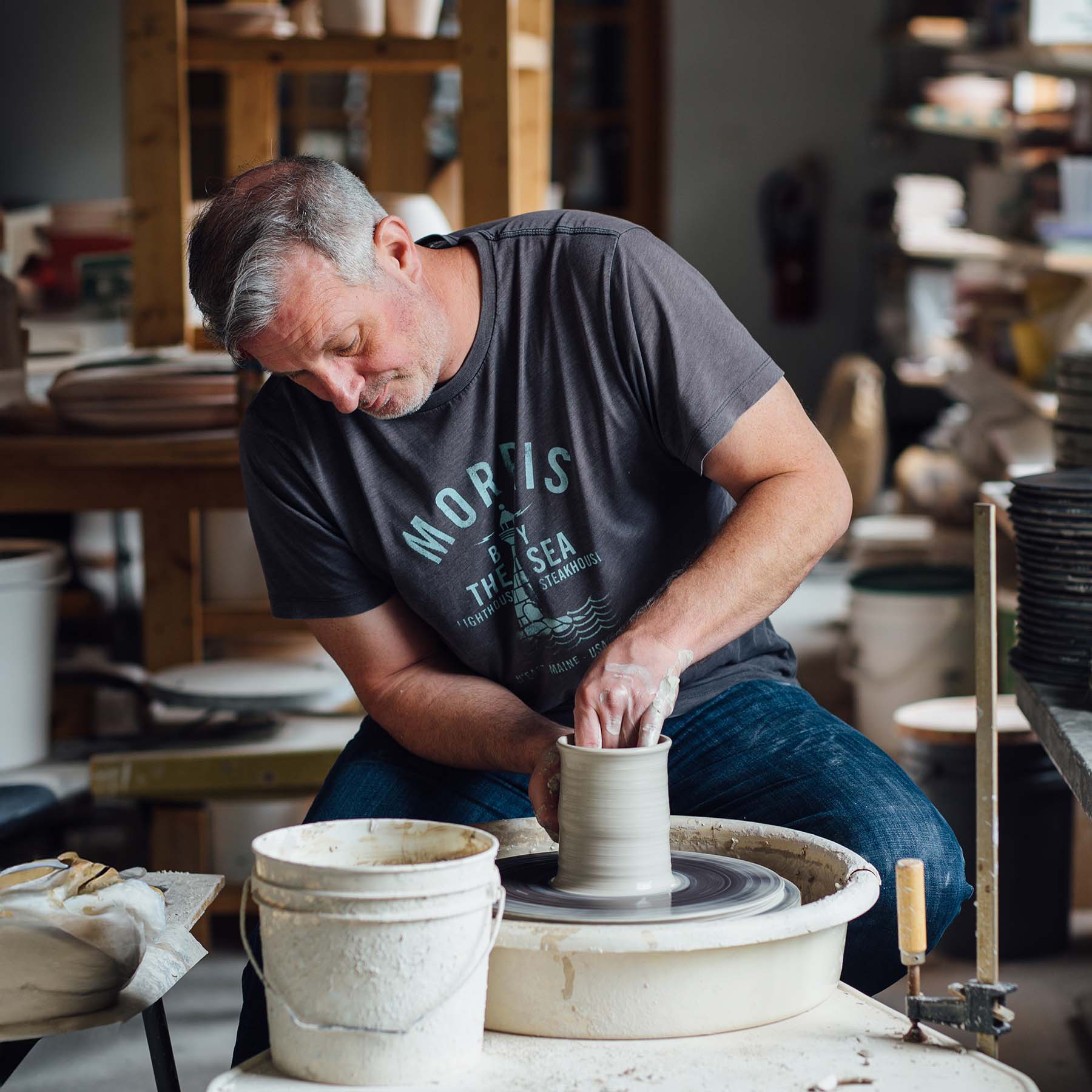 Hamilton Williams sitting at a potter's wheel while shaping spinning clay into a cylinder.