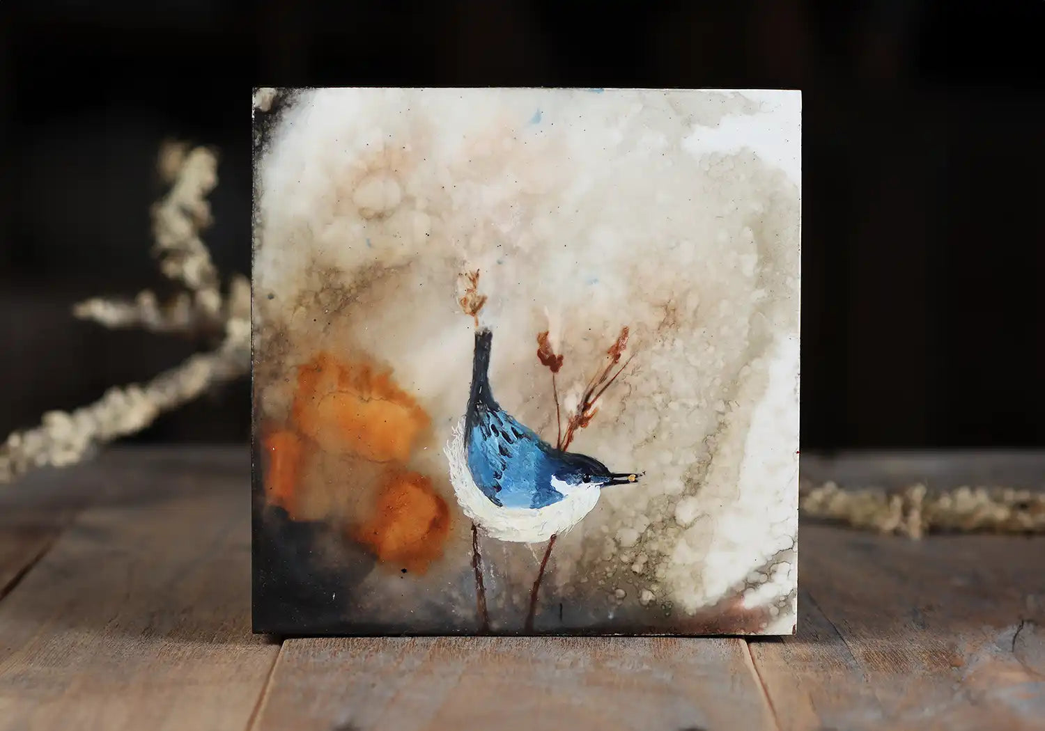 An small square encaustic painting that depicts a blue bird alighting on a limb, a naturalistic background with taupes and oranges.