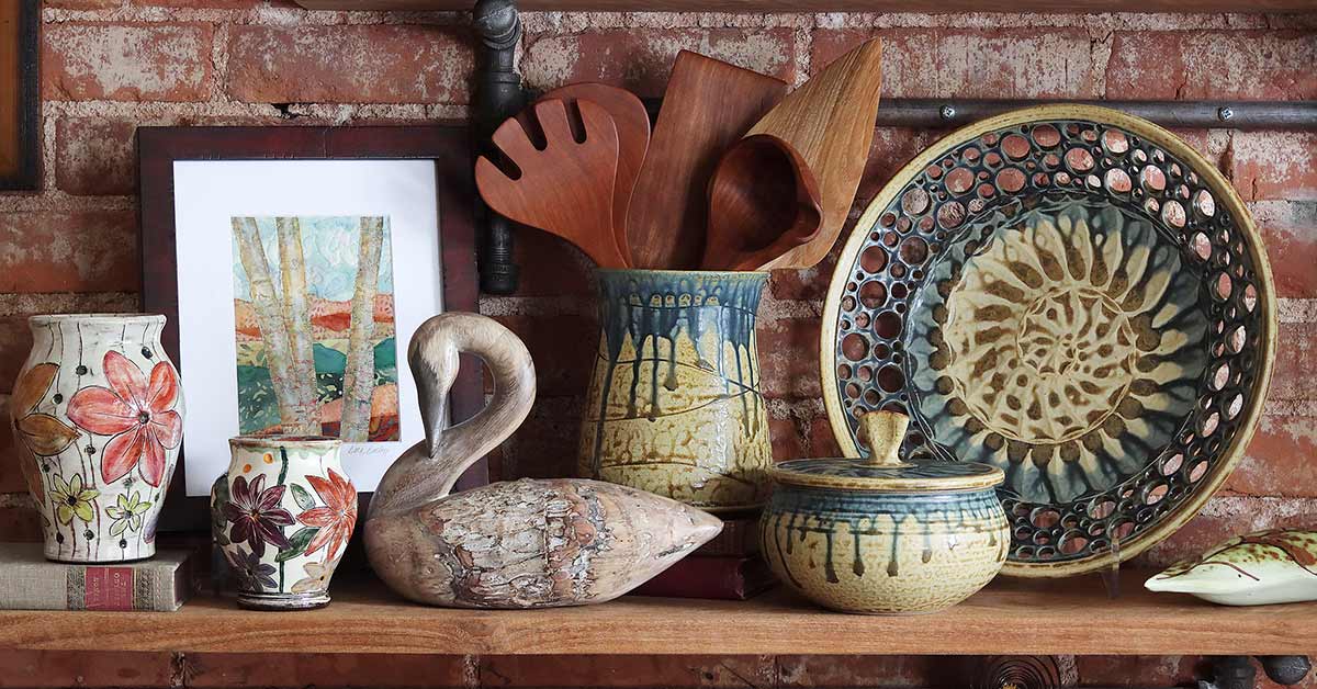 An assortment of pottery, quilting, wooden utensils, and a wooden swan decoy rest on a shelf against a brick wall.