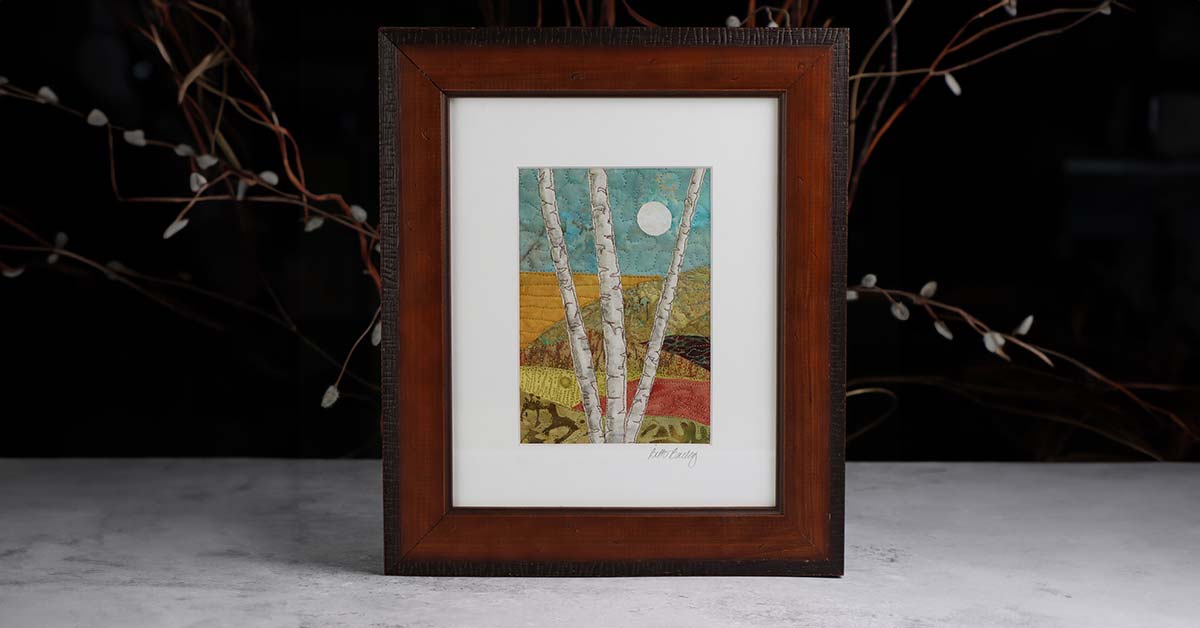 A wood-framed piece of fiber art by Beth Bailey that depicts two birch trees against a colorful landscape, all rendered in quilted fabrics.