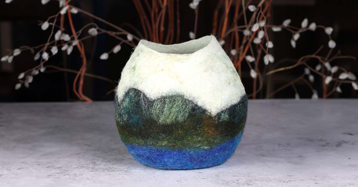 A felted vessel that depicts a landscape with mountains and a lake in the foreground.