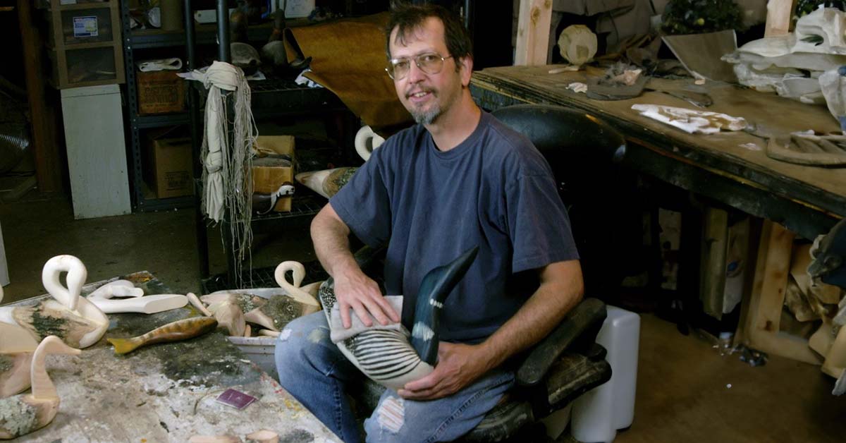 Avian decoy-maker, Chris Boone, sitting in his wood studio holding one of his completed mallard duck decoys while wood components of decoys-in-progress surround him.