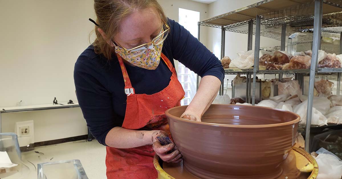 A woman, Courtney Long, wearing a mask and a studio apron, leaning over a large bowl of soft clay as she shapes it on a potter's wheel.