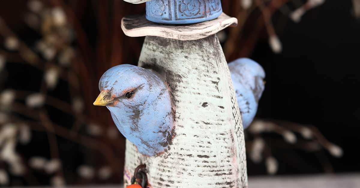 A close view of a ceramic sculpture consisting of decorative clay pods stacked like a totem. The close up is of a pod with two bluebird heads rendered.