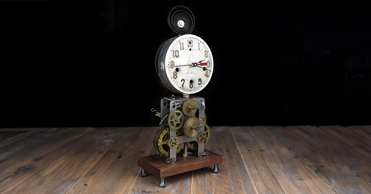 An eclectic clock made by Carol Louise Mayer shows open gears and a ticking feather second hand as it sets alone on a wooden table with a black background.