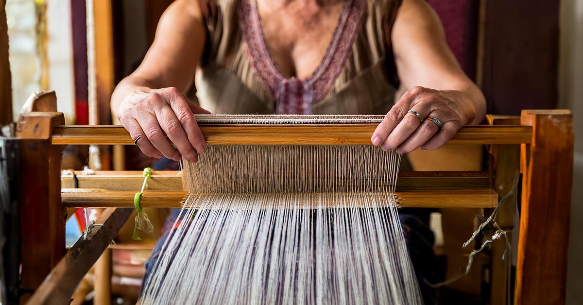 A weaver's hands grasp the heddle of a loom that is strung with warp fibers. 