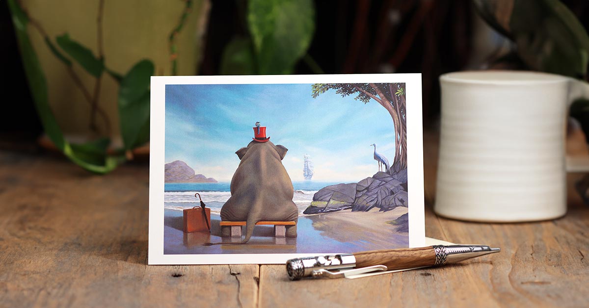 A small vignette consisting of a greeting card with an image of a anthropomorphize elephant sitting on a beach on the front setting upright on a wooden surface, a wooden pen, a white mug, and a potted plant disappearing into the background.