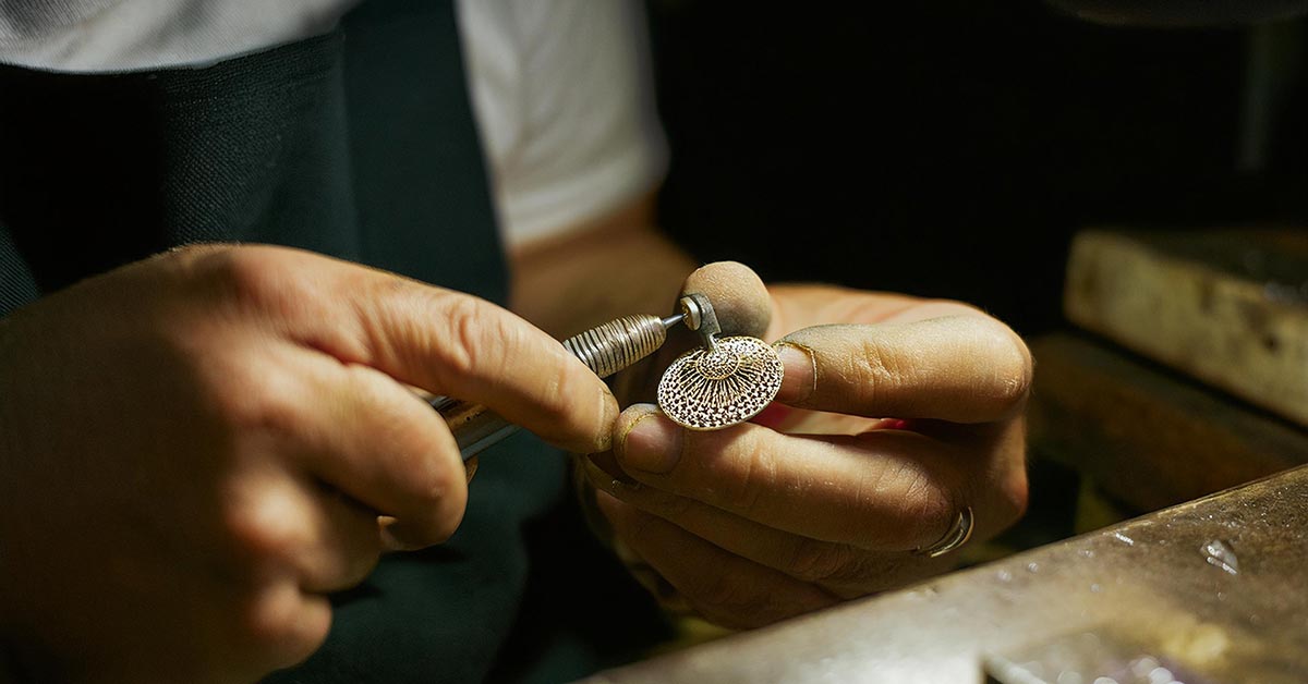 A jewelry-maker's hands polish a brooch in a dimly light workshop.