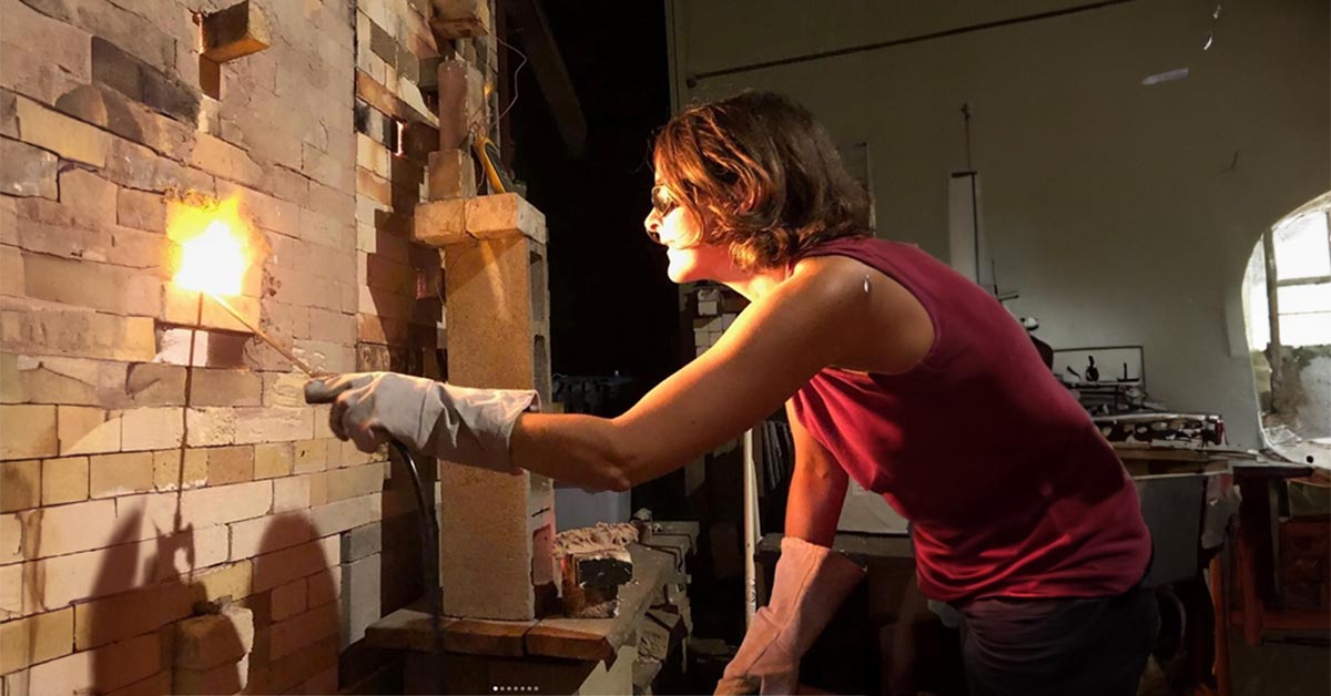 A woman, Joy Tanner, leaning over to peer into a fiery kiln.