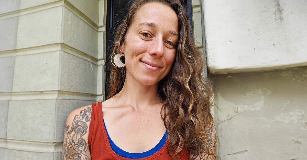 A young woman, Laurin Jennings, leans against the corner of building with a doorway behind her smiling into the camera.