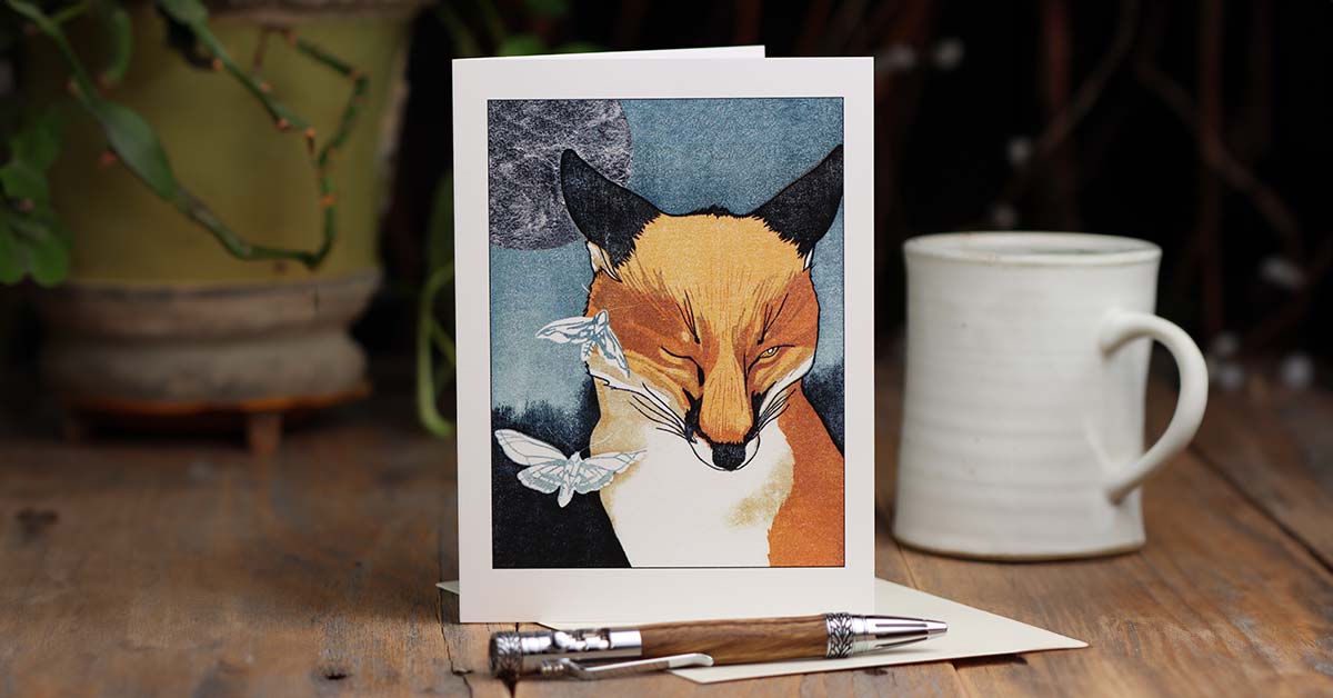 A small vignette consisting of a greeting card with an image of a winking fox on the front setting upright on a wooden surface, a wooden pen, a white mug, and a potted plant disappearing into the background.