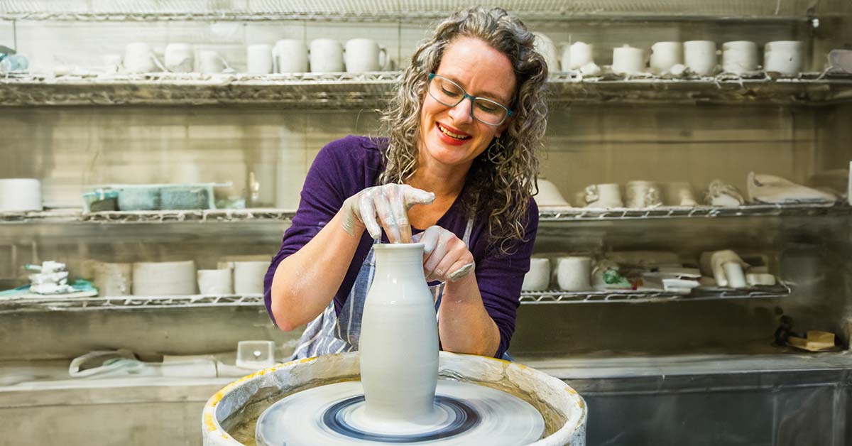 A woman with glasses and long hair, pleasantly smiling and gently finishing the lip of a freshly thrown vase on a potter's wheel. Shelves of additional pottery in-progress are in the background.