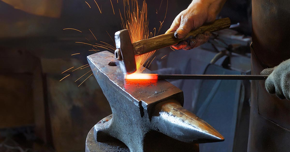 The hands of a apron-clad blacksmith hold a piece of iron with a glowing hot tip against an anvil as a hammer rings down, sparks flying.