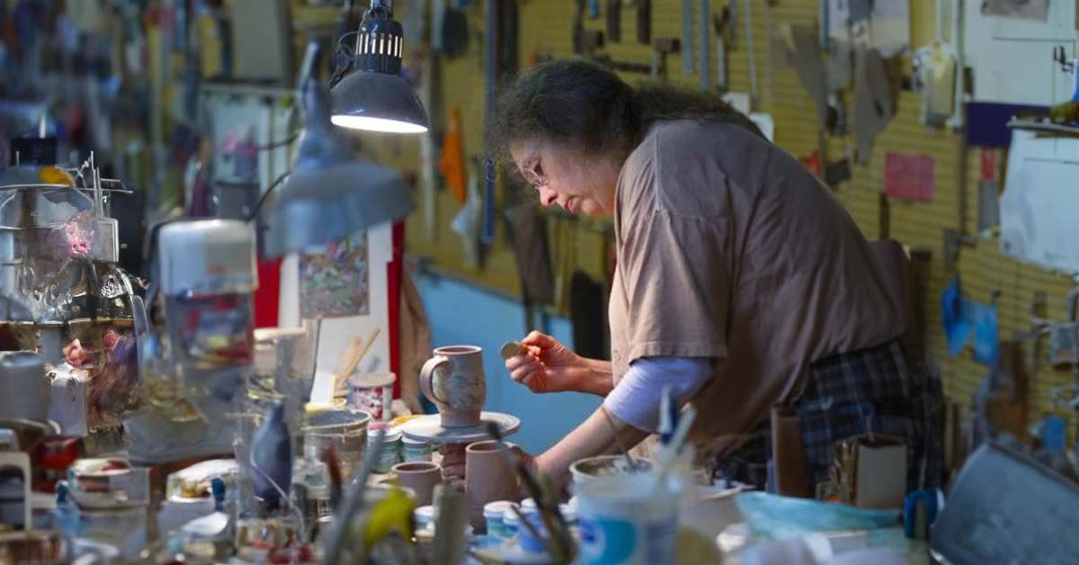 A woman ceramics artist, Molly Lithgo, leaning over with an artist's brush to decorate a handmade mug in her studio.