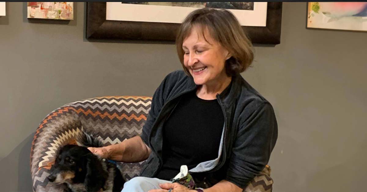 Encaustic artist, Nadine Koski, sitting on a chair petting a small dog. There is framed art on the wall behind her.