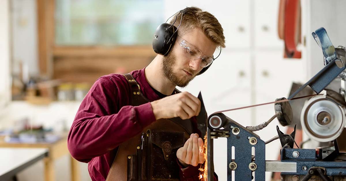 A young man, Raleigh Avery, wearing safety glasses and ear protection, grinding a forged knife blade into its final shape using a belt sander.