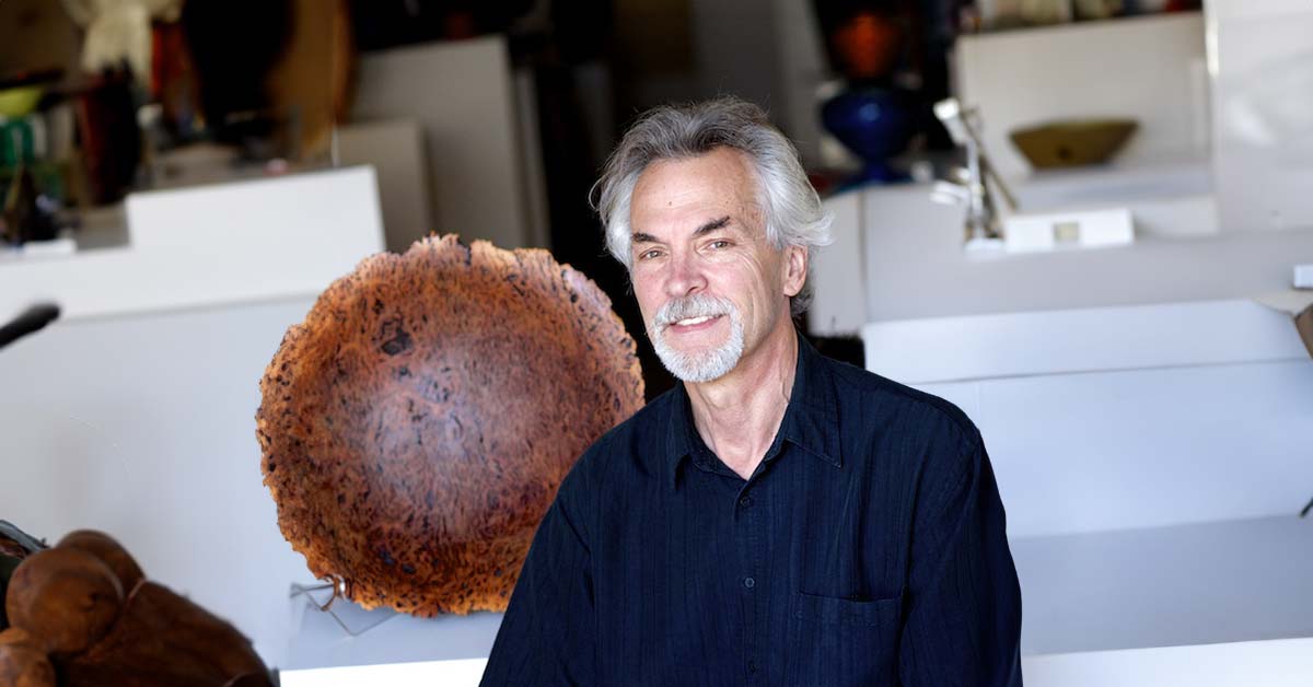 An older man with a goatee and mustache, Steve Noggle, sitting in front of one of his wood-turned bowls.