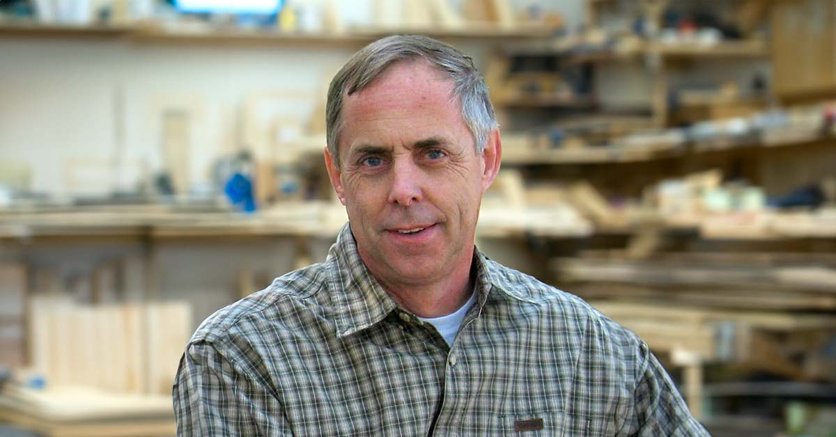 An older man, woodturner Warren Carpenter, facing the viewer while sitting in a woodworking studio.